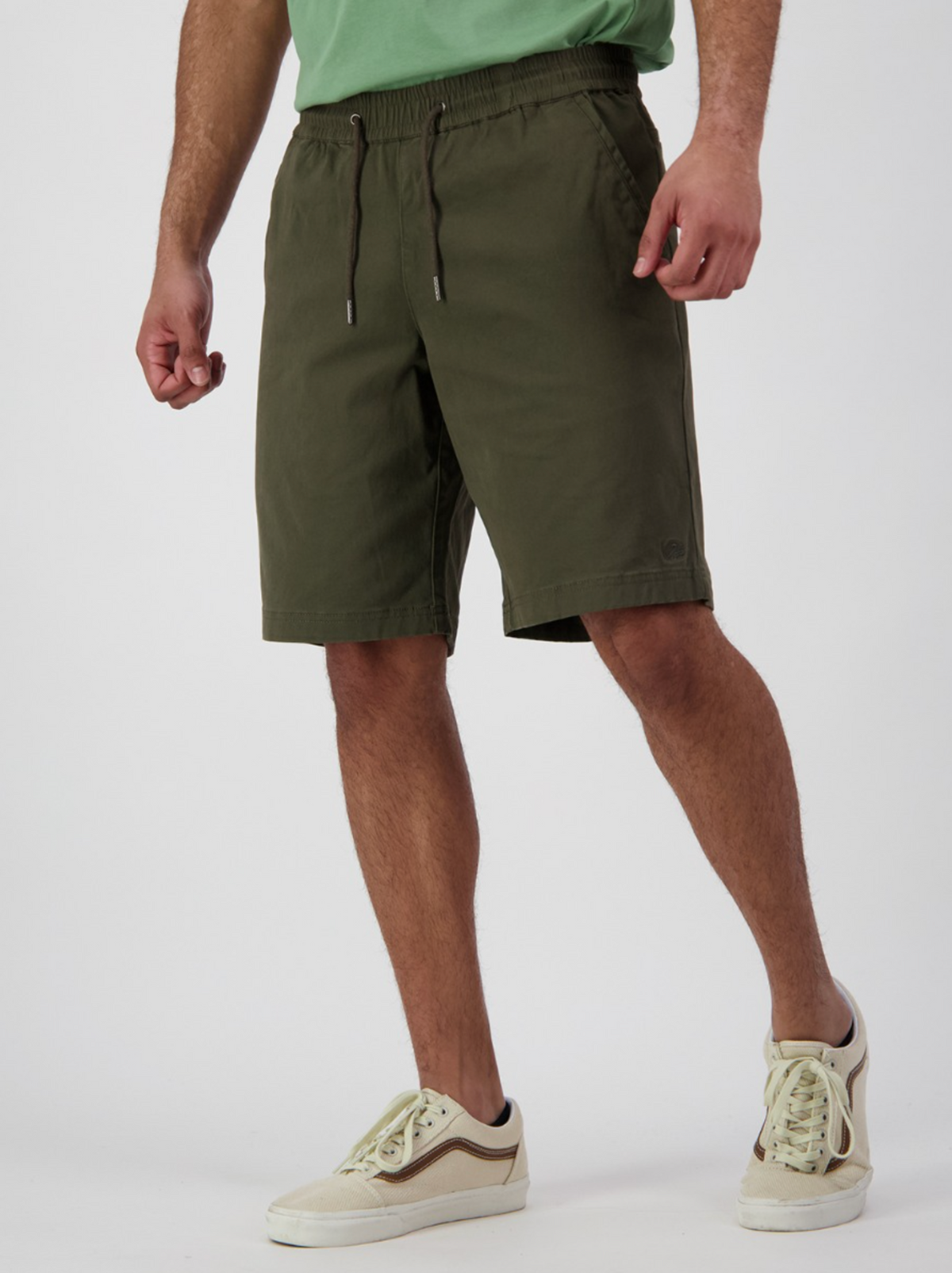 A person in Swanndri Clevedon olive green stretch canvas shorts and a light green shirt stands with their hands relaxed at their sides, wearing white sneakers with brown accents against a plain white background.