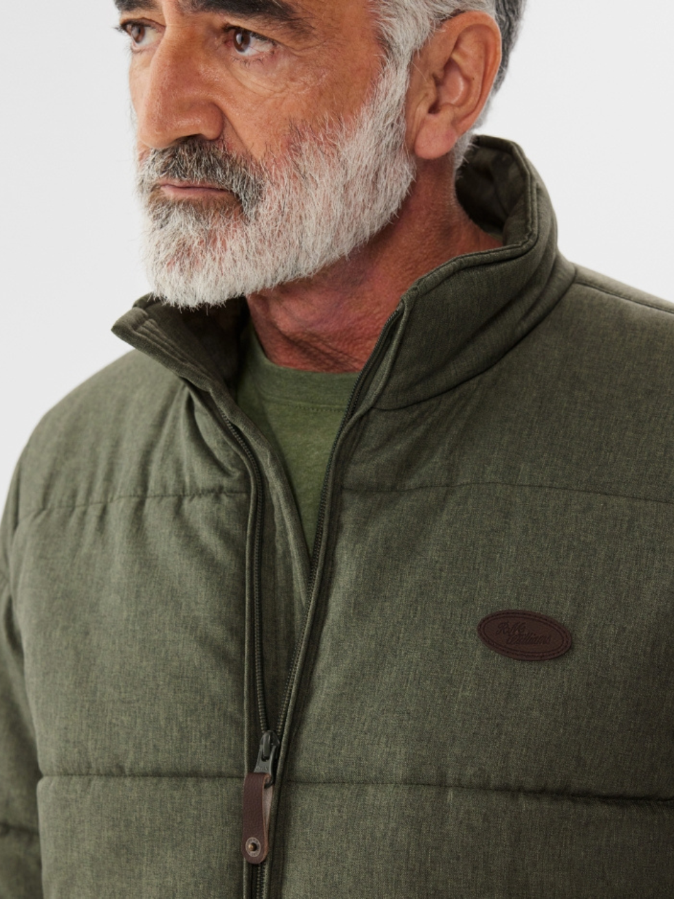 An older bearded man wears a green RM Williams Patterson Creek Jacket, a quilted puffer with a high collar and partially open zipper revealing a green shirt underneath. A small oval logo adorns the chest of this water-resistant jacket against a plain white background.