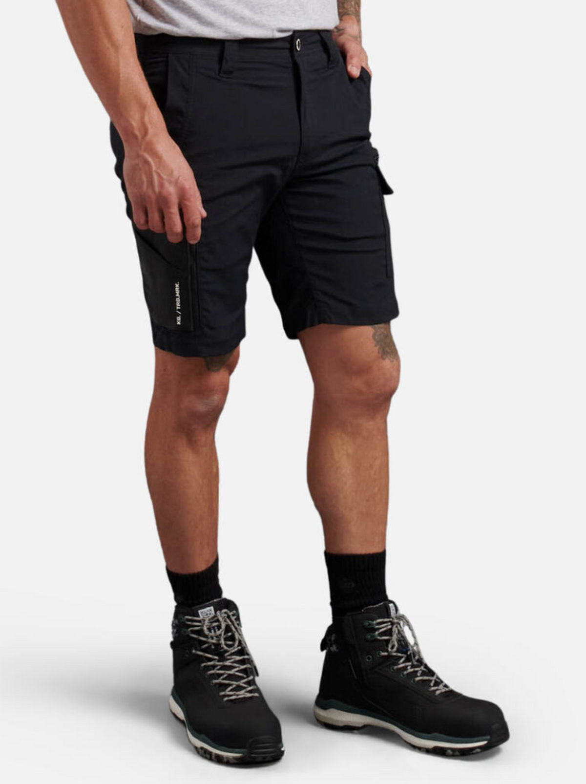 Someone is shown from the waist down against a plain light background, wearing black King Gee Trademark Cargo Shorts made from stretch ripstop fabric, black socks, black hiking boots with white laces, and a light gray t-shirt.
