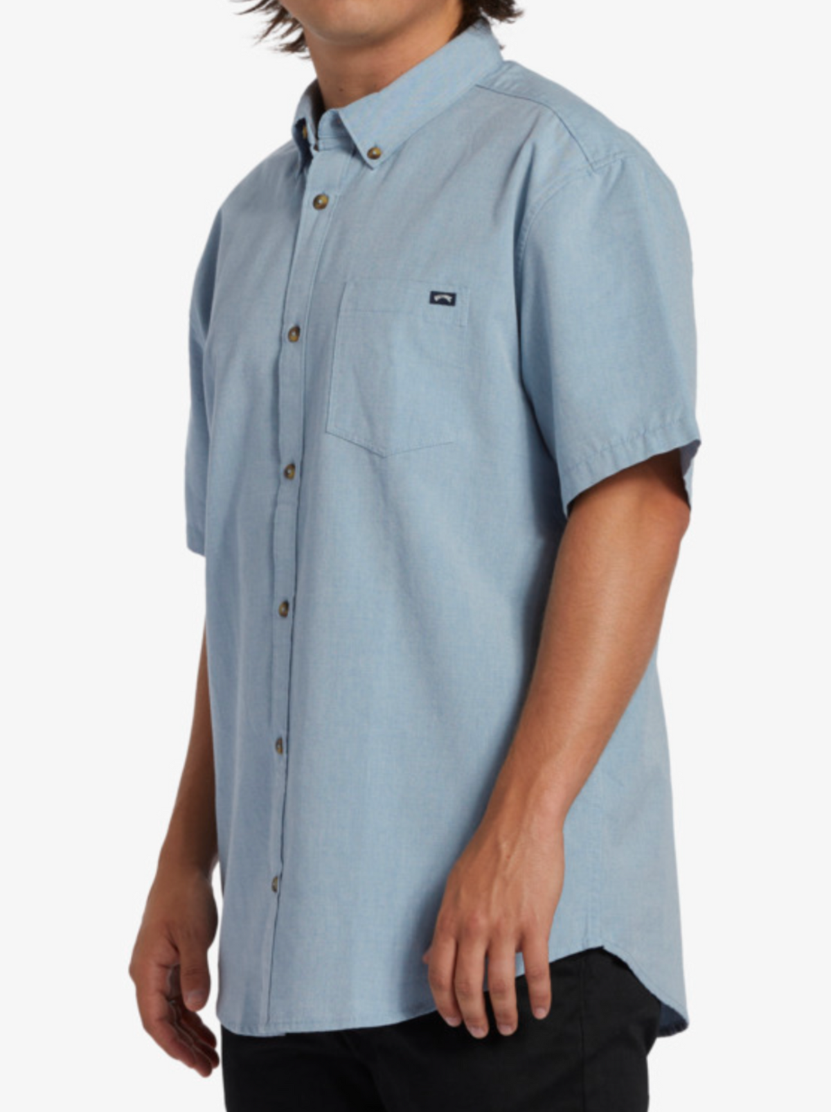 A person showcases modern style and classic roots in the Billabong All Day Short Sleeve Shirt, featuring light blue woven fabric and a chest pocket, paired with black pants against a plain white background.