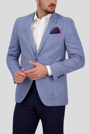 A man is dressed in a Bruton Asher Linen Blazer in light blue, paired with a white dress shirt and dark trousers. A red pocket square adorns the breast pocket of the professional formal blazer. With his hands positioned in front of him, he gazes slightly to the side against a plain gray background.