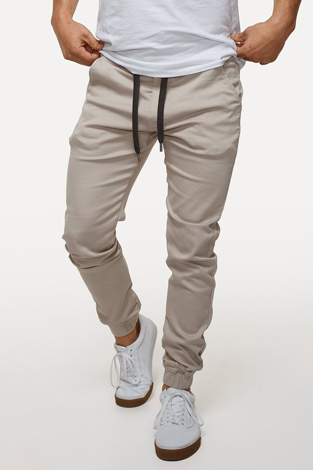 A person confidently poses with hands in pockets, dressed in beige Industrie The Drifter Chino Pants with an elastic waist and white sneakers with black laces. Their crisp white t-shirt is neatly tucked into the chinos, while a minimalist all-white background complements the ensemble.