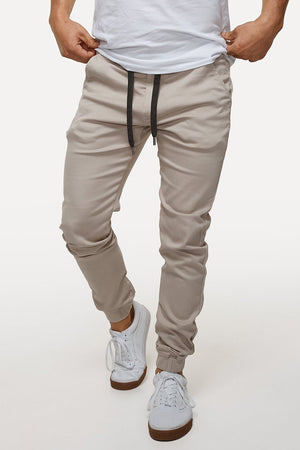 A person confidently poses with hands in pockets, dressed in beige Industrie The Drifter Chino Pants with an elastic waist and white sneakers with black laces. Their crisp white t-shirt is neatly tucked into the chinos, while a minimalist all-white background complements the ensemble.