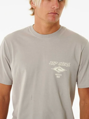 A young man with blonde hair models the Rip Curl Fade Out Icon Tee, a light grey cotton t-shirt with a small white logo and text on the left chest, standing against a plain white background.