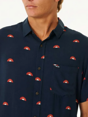 A person wearing the Rip Curl Party Pack Short Sleeve Shirt in navy blue with a pattern of small red and white semi-circles stands against a plain light background, showing the upper body and part of the face, highlighting its comfortable fit.