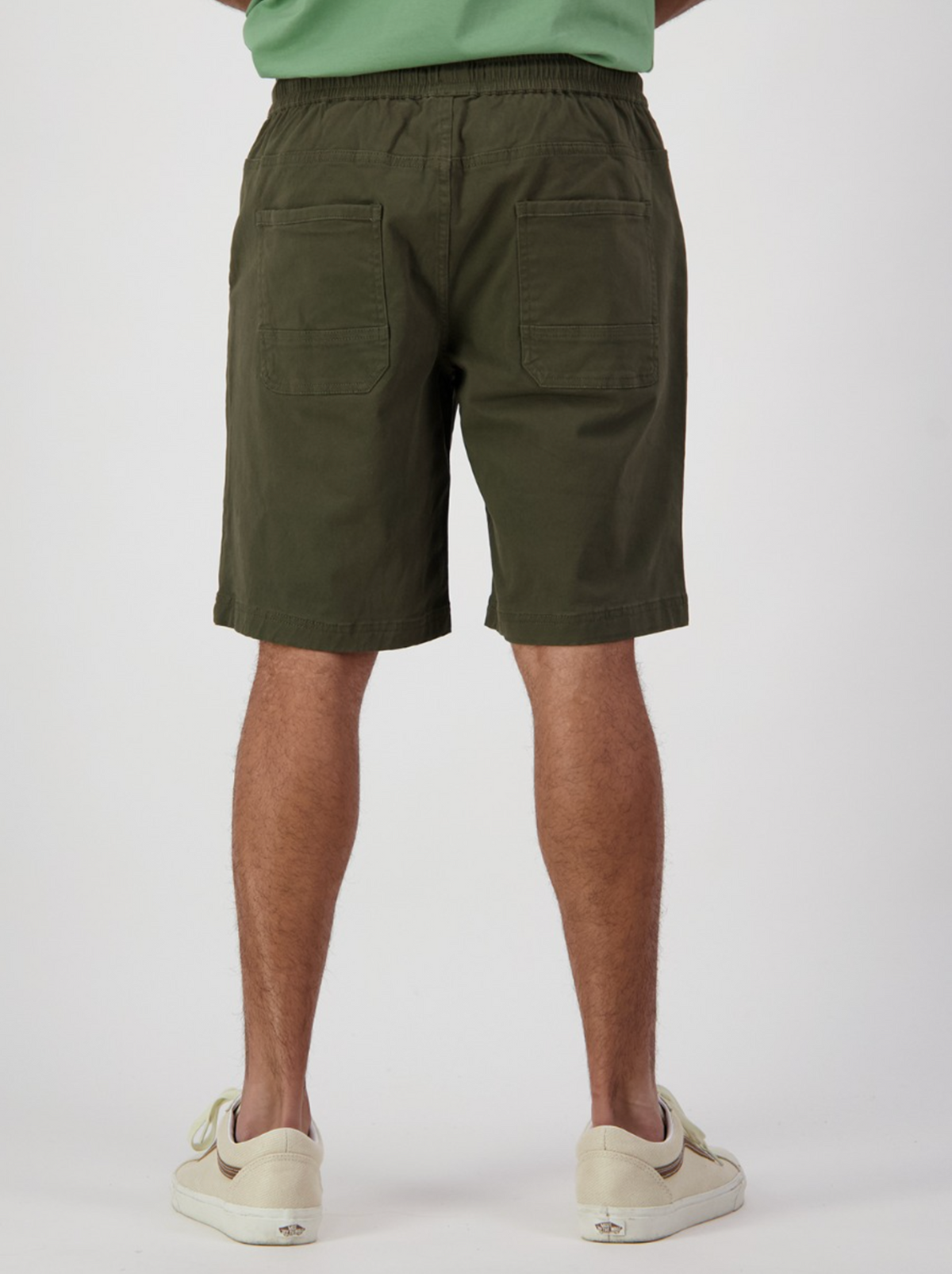 A person faces away from the camera in Swanndri Clevedon Stretch Canvas Shorts, showcasing two back pockets. Paired with white sneakers and a matching green shirt, this outfit stands out against a plain white backdrop, emphasizing the relaxed comfort of elastic waist shorts.