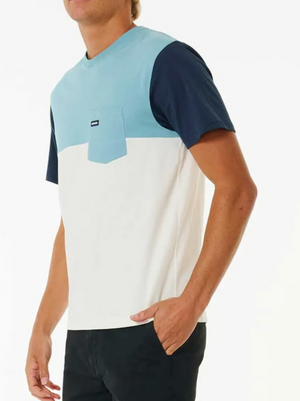 A person is seen wearing a Rip Curl Surf Revival Custom Tee, featuring light blue, navy, and white color blocks with a small chest pocket. This 100% cotton tee is paired with black pants; one hand rests in their pocket against a plain white background.