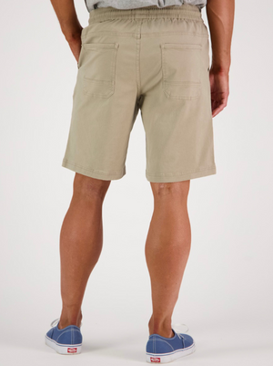 A person wearing Swanndri Clevedon Stretch Canvas Shorts in beige, featuring an elastic waist and back pockets, is paired with blue slip-on sneakers. The scene is set against a plain white background.