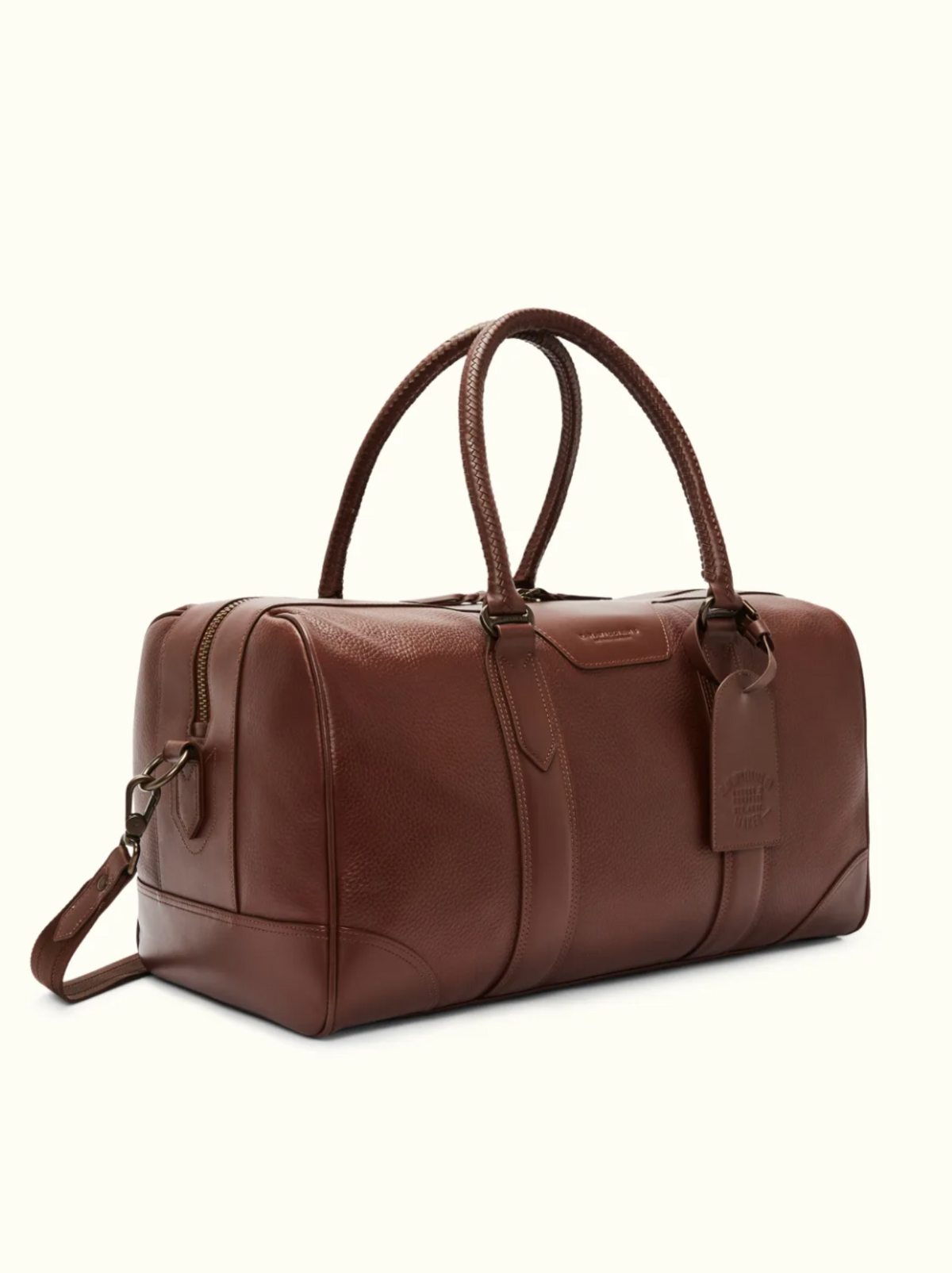 The RM Williams Overnight Bag is a brown full-grain leather duffel with dual handles, a detachable shoulder strap, and sleek metal hardware. Ideal for travel, it features a luggage tag and embossed branding on the side. Displayed on a neutral background.