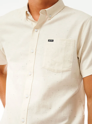 Someone wears the Rip Curl Ourtime Short Sleeve Shirt in light beige, featuring a chest pocket with a small black label above it and a subtle speckled pattern. Made from 100% cotton herringbone. The person's head is not visible.