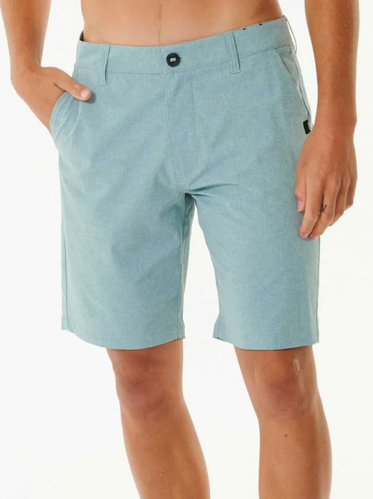 A person wearing the Rip Curl Boardwalk Phase Nineteen Short in light blue, featuring a button and zipper, knee-length fit, with one hand in the pocket, stands against a plain white background.