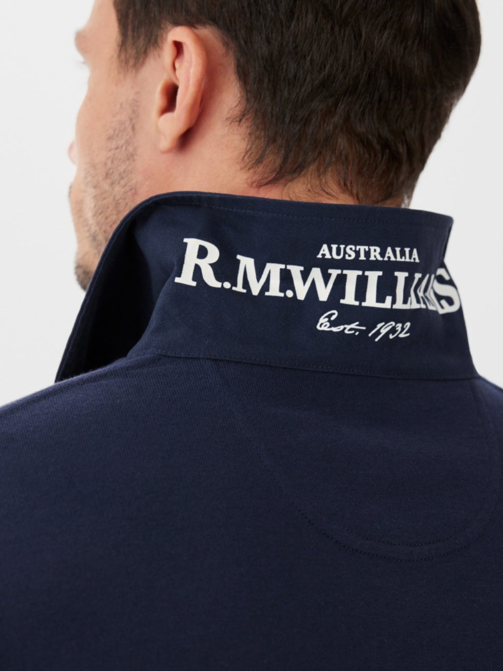 A person seen from behind wears the RM Williams Tweedale Rugby, a navy heavyweight collared shirt with "R.M. Williams Australia Est. 1932" printed in white on the turned-up collar.