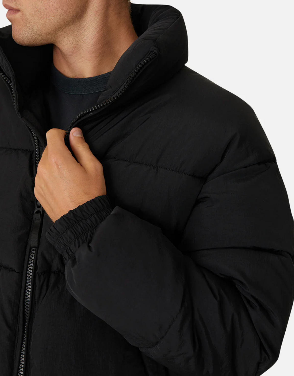A person wears the Industrie The Regent Puffer Jacket, a black puffer with quilted padding, pulling up its front zipper. This jacket features a high collar and elastic cuffs against a plain white background.