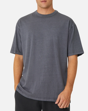 A person wears a loose-fitting, short-sleeved Industrie The Del Sur Tee in dark gray with black pants against a plain background. This 100% cotton crew neck T-shirt features an oversized, slightly faded casual look. The person's face is not visible.