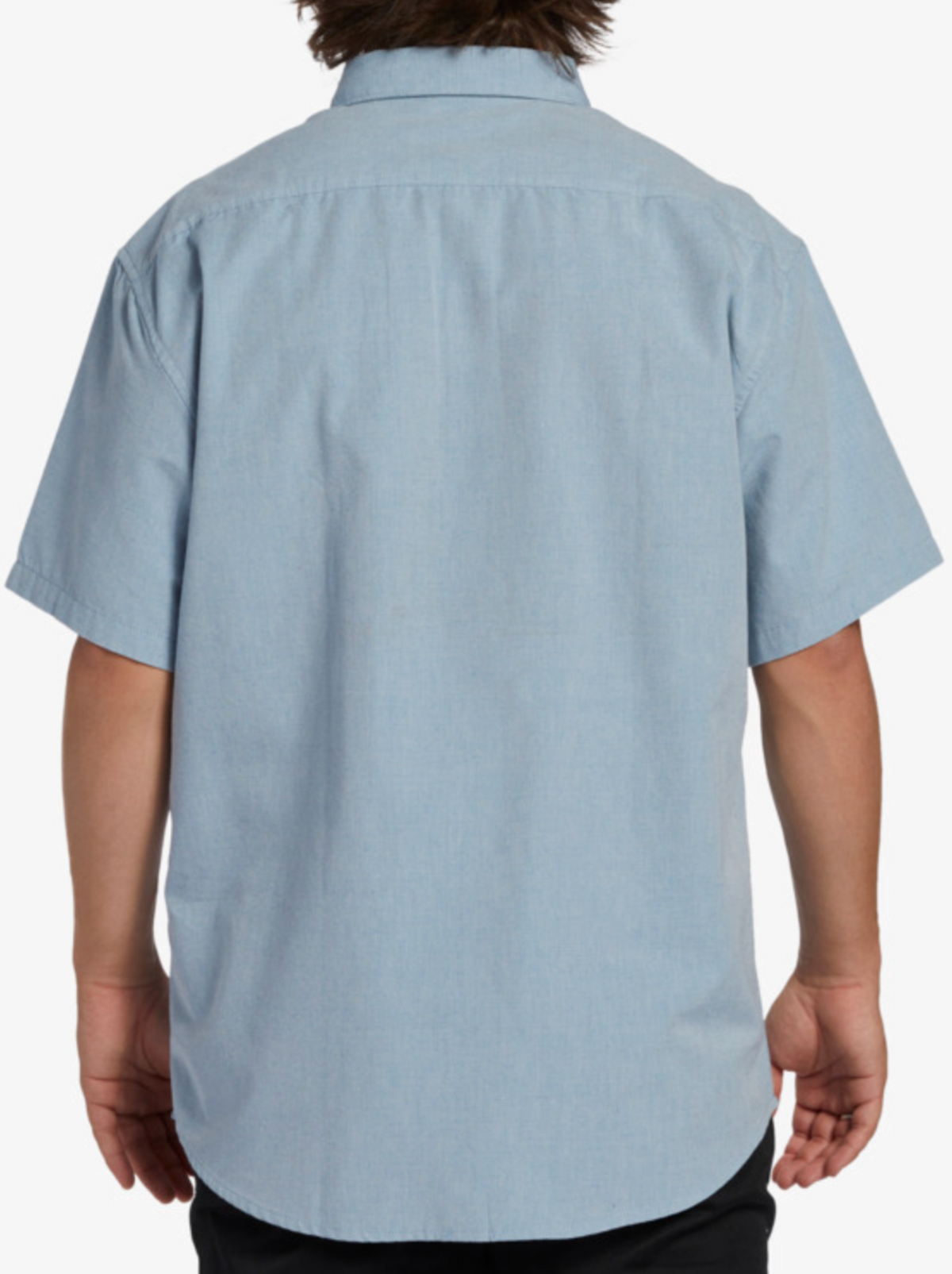 A person is shown from the back wearing a light blue Billabong All Day Short Sleeve Shirt made from woven fabric with a simple collar and dark pants; the shirt has no visible patterns or logos, embracing classic style.