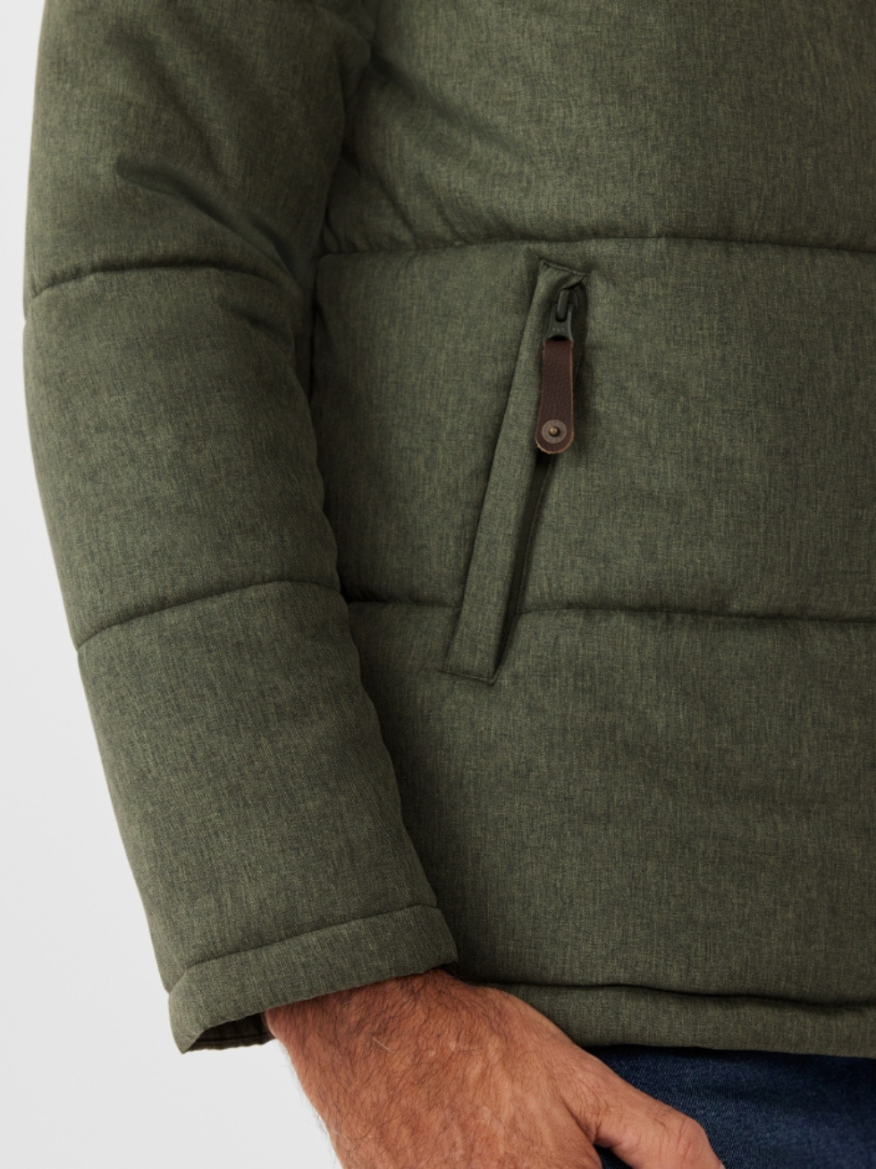 A person sports the RM Williams Patterson Creek Jacket, a green quilted puffer with a zippered pocket and water-resistant textured fabric, paired with blue pants. One hand is visible.