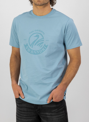 A man stands with his hands in his pockets, wearing a light blue Swanndri Original v2 Print T-Shirt with a circular chest logo and black jeans, set against a plain white background.