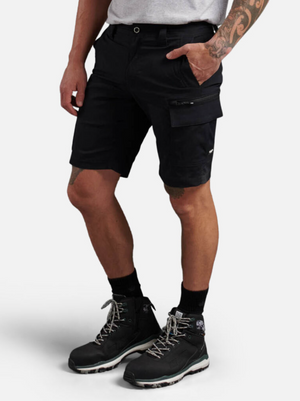 A person wears black King Gee Trademark Cargo Shorts, a light gray T-shirt, black socks, and black hiking boots. One hand is in a pocket, showing arm tattoos. The background is plain and light-colored.