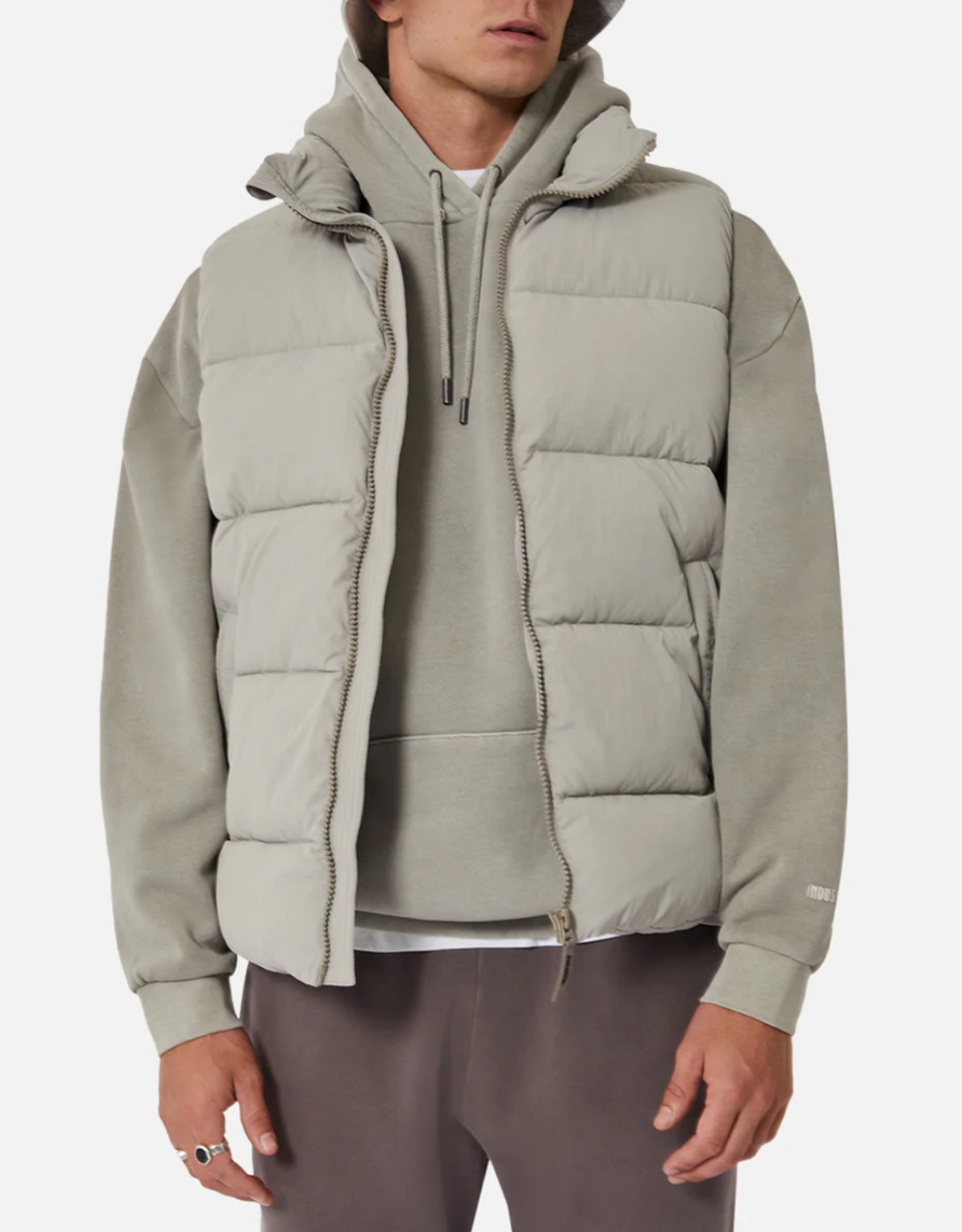 A person is wearing the Industrie The New Hanam Puffer Vest in light grey with thick quilted padding over a grey hoodie. They have on darker grey pants and a ring on their left hand. Their face is not visible, and the background is plain white.