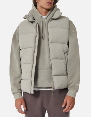 A person is wearing the Industrie The New Hanam Puffer Vest in light grey with thick quilted padding over a grey hoodie. They have on darker grey pants and a ring on their left hand. Their face is not visible, and the background is plain white.