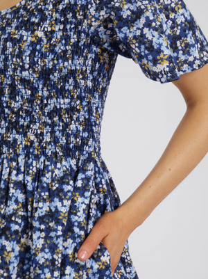 A close-up of a person wearing the Foxwood Lulu Dress in sapphire ditsy print, made from fresh cotton poplin with a gathered ruched bodice and their right hand casually placed in a pocket.