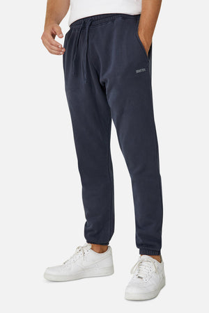 A person wearing Industrie The Del Sur Track Pant in navy blue with an elastic drawstring waist and small Industrie logo on the upper thigh, paired with white sneakers, stands against a plain white background.