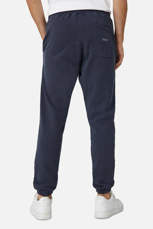 A person in Industrie The Del Sur Track Pant, featuring a navy blue color, elastic drawstring waist, back pocket, elastic cuffs, and an embroidered logo, stands with their back to the camera. They're paired with a white shirt and sneakers against a plain white background.