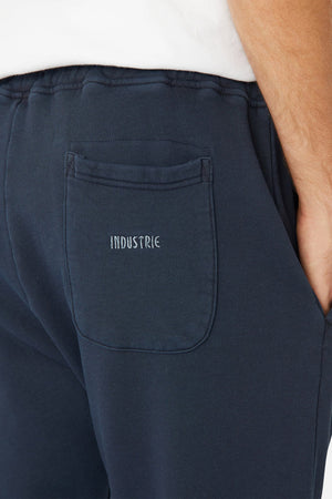 A person wears the Industrie The Del Sur Track Pant in navy, showcasing an embroidered white logo on the back pocket. Their hand is partially visible, casually tucked into the pocket.