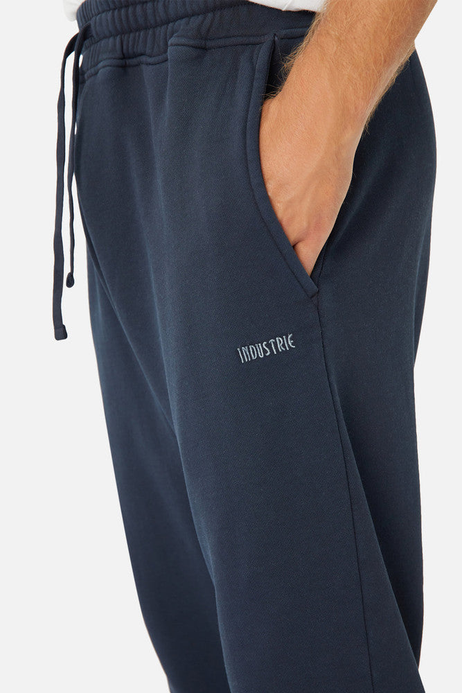 A close-up shows someone in Industrie The Del Sur Track Pant, featuring an elastic drawstring waist and the person's hand resting casually in a pocket. The embroidered Industrie logo enhances its stylish appeal.