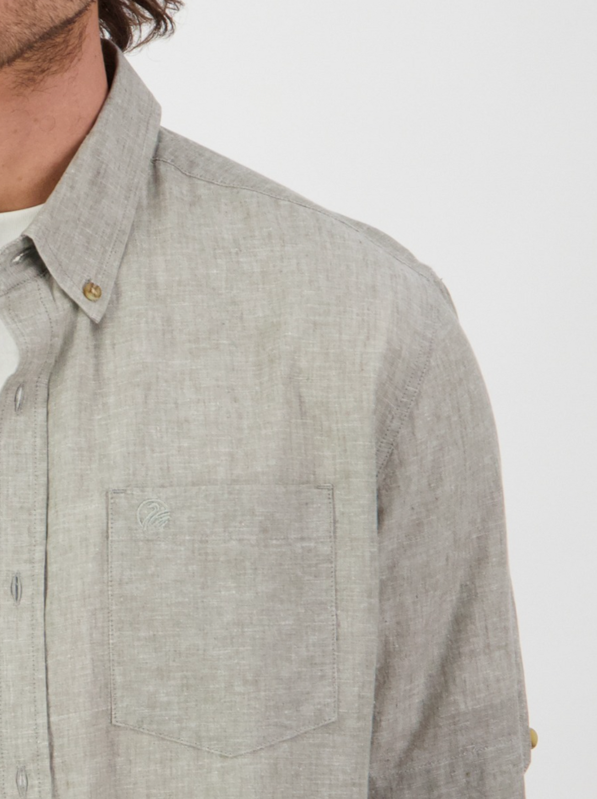 Someone in a light gray Swanndri Lowell v2 Short Sleeve Shirt with an embroidered swan logo stands against a plain background. The focus is on the upper half of the linen-cotton blend shirt, highlighting its texture and buttons, while only part of their face is visible.