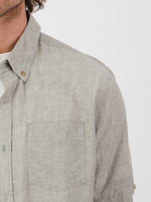 Someone in a light gray Swanndri Lowell v2 Short Sleeve Shirt with an embroidered swan logo stands against a plain background. The focus is on the upper half of the linen-cotton blend shirt, highlighting its texture and buttons, while only part of their face is visible.