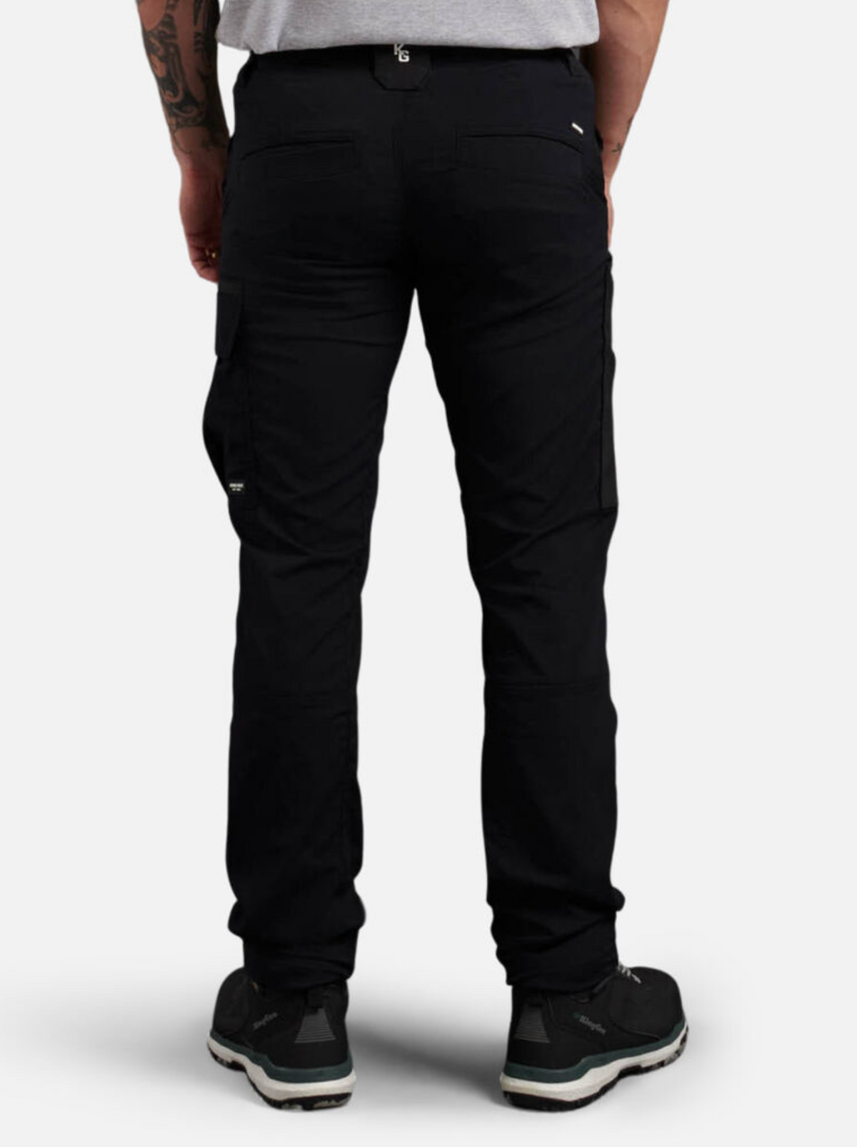 A person wearing black King Gee Trademark Cargo Pant, a light gray t-shirt, and black sneakers stands with their back to the camera, displaying the rear view of the pants. A tattoo is visible on their left forearm.