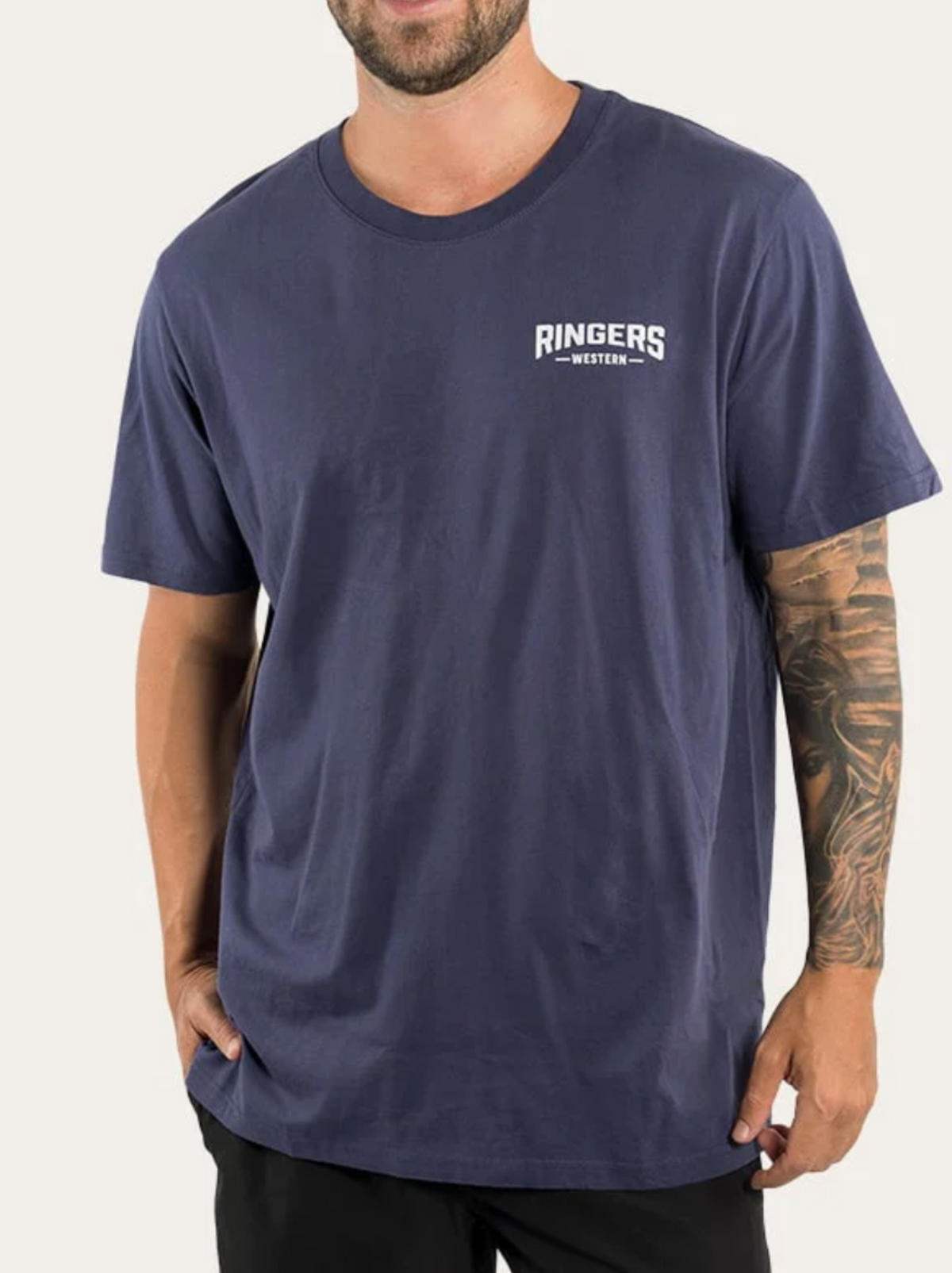 A man wears the Ringers Western Squadron Loose Fit T-Shirt in navy blue, made from 100% cotton jersey with short sleeves and a small white chest logo, paired with black pants. He has a tattoo sleeve on his right arm and his left hand in his pocket.