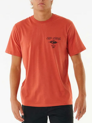 A person wears the Rip Curl Fade Out Icon Tee, an orange 100% cotton short-sleeve t-shirt with a small black "Rip Curl" logo and text on the left chest, standing against a white background.