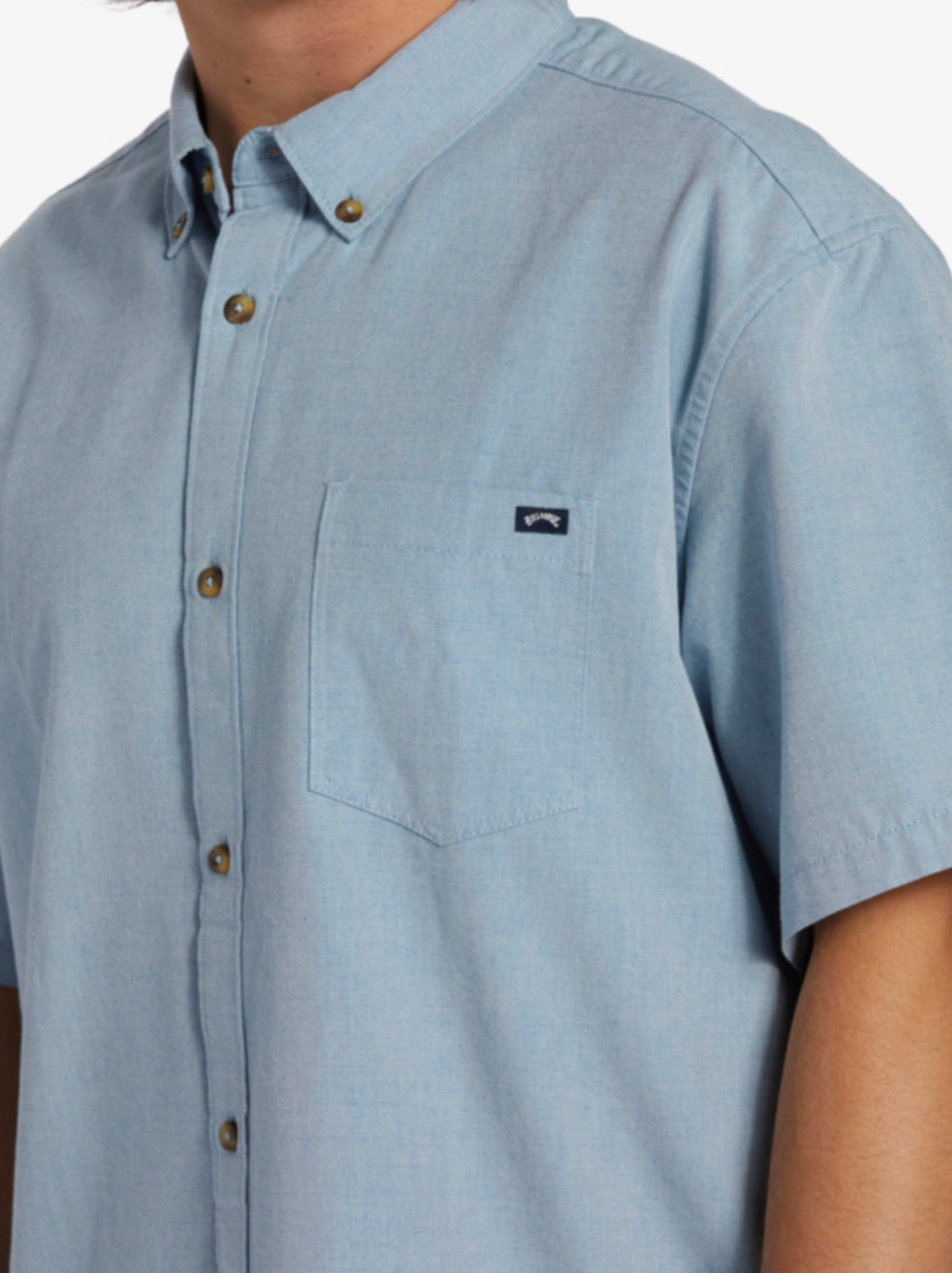 A person wears the Billabong All Day Short Sleeve Shirt in light blue woven fabric, short sleeves, brown buttons, and a chest pocket with a small rectangular logo. Their head is partially out of frame. Classic Roots style meets modern comfort.