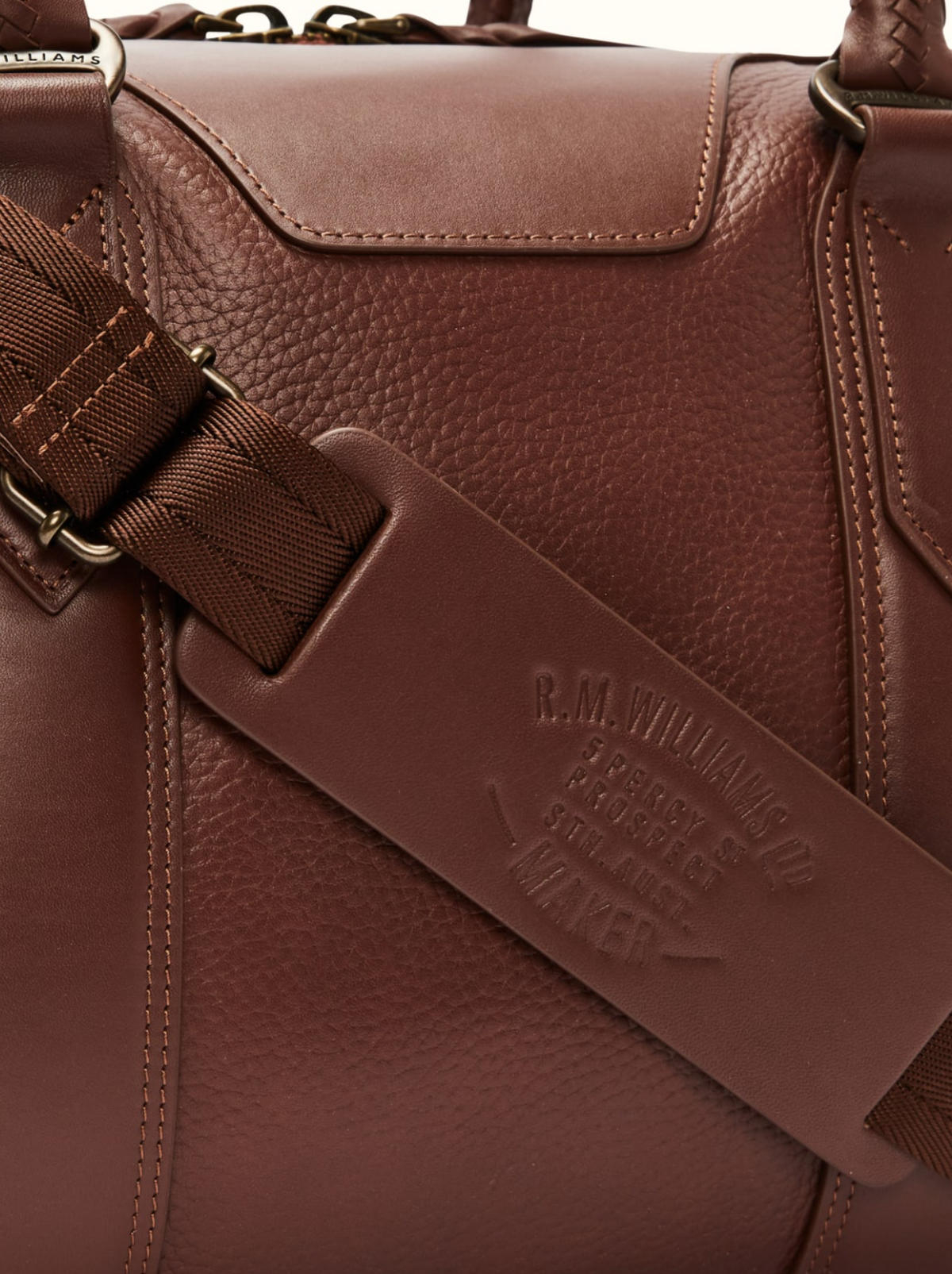 Close-up of the RM Williams Overnight Bag in brown full grain leather, showcasing detailed texture and a brown strap with metal hardware. The embossed strap features "R.M. Williams," "leather," and "Australia," making it an exceptional choice for travel items like an overnight bag.