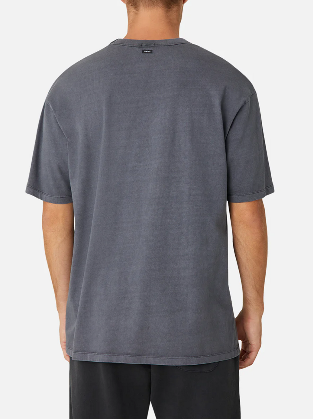 A person is shown from the back against a plain background, wearing an oversized, faded black Industrie The Del Sur Tee and black pants. The T-shirt features short sleeves, 100% cotton fabric, and a relaxed silhouette.