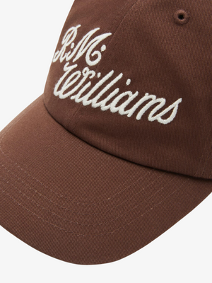 A close-up of the RM Williams Script Cap, a brown cotton twill baseball cap featuring white "R.M.Williams" script embroidery on the front.