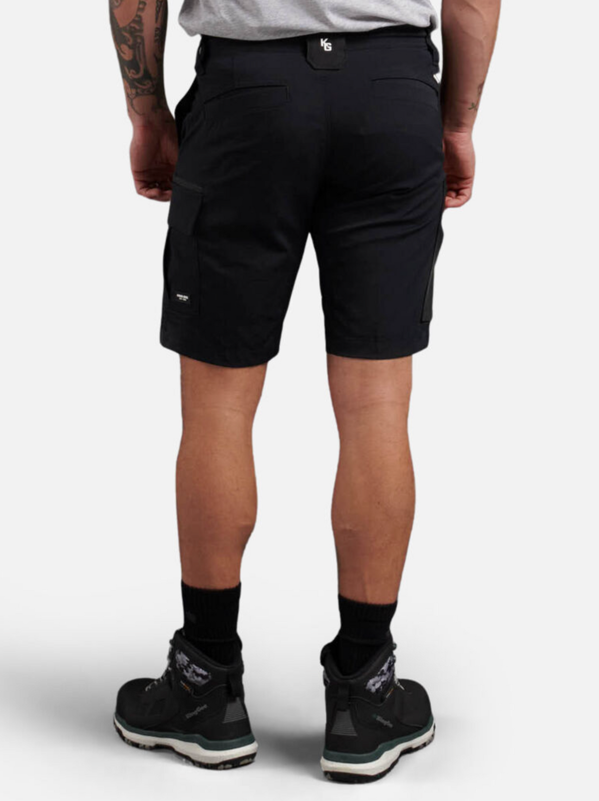 A person is shown from behind wearing King Gee Trademark Cargo Shorts in black and black athletic shoes, with a tattoo visible on their left arm, standing against a plain white background.