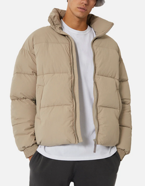 A person wearing Industrie The Regent Puffer Jacket in beige with quilted padding over a white t-shirt and black pants. They are also sporting a light grey bucket hat. The jacket, featuring an Industrie label, is unzipped, and the person's hands rest in their pant pockets against a plain white background.