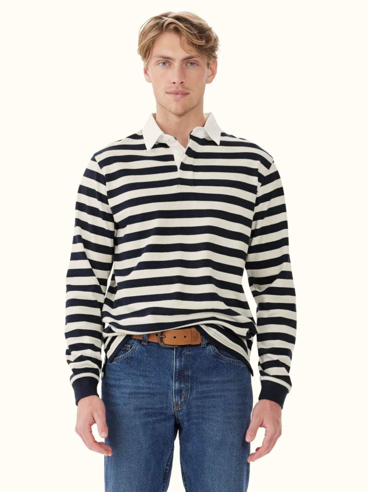 A man stands facing forward against a light background, wearing the RM Williams Tweedale Rugby—a heavyweight black and white horizontal striped jersey with a cotton twill collar—paired with blue jeans and a tan belt.
