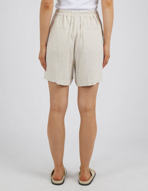 A person is seen from behind wearing Foxwood Farrow Shorts featuring an elastic waistband and two pockets. They are matched with a white shirt over a neutral backdrop, accessorized with sandals for a comfy fit.