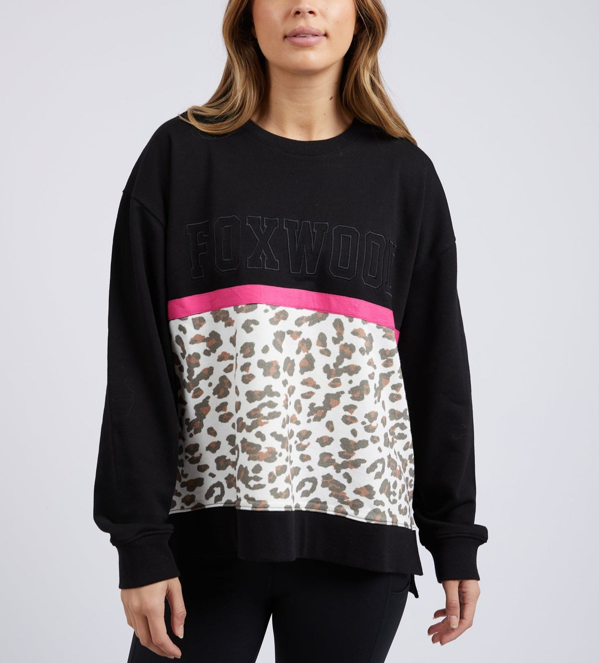 A woman models the Foxwood Split Crew, a black sweater featuring "FOXWOOD" in bold text, a vibrant pink stripe, and leopard print detailβan ideal addition to any casual wardrobe.