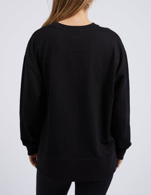 A person with long light brown hair stands with their back to the camera, wearing the relaxed Foxwood Split Crew sweater and black pantsβan ideal piece for any casual wardrobeβagainst a plain light background.