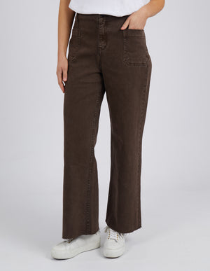 A person stands confidently in the Foxwood Eddie Wide Leg Jean, featuring large front pockets and raw edge detailing, paired with a crisp white shirt and sneakers. The plain light gray background complements this effortlessly stylish look.