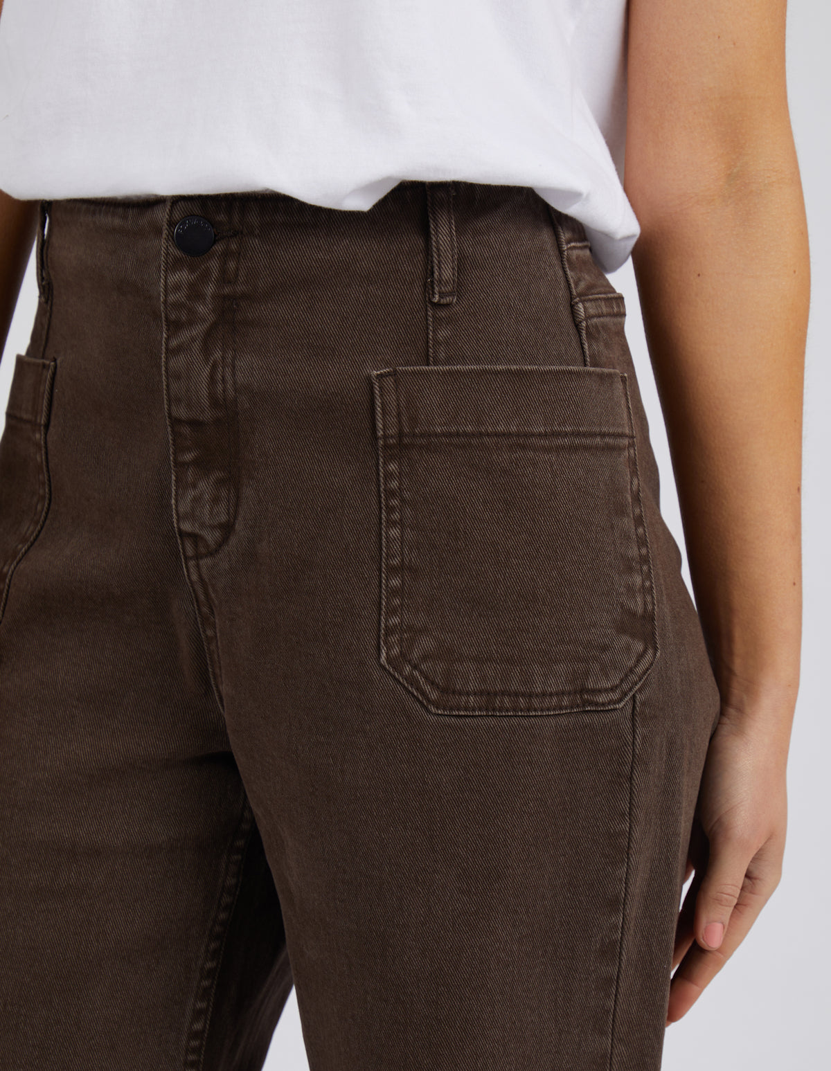 A person models Foxwood Eddie Wide Leg Jean in brown, paired with a white shirt. The image focuses on the midsection, highlighting the jeans' high-waisted fit, wide legs, large front pockets, and subtle raw edge detailing.