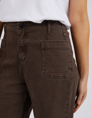 A person models Foxwood Eddie Wide Leg Jean in brown, paired with a white shirt. The image focuses on the midsection, highlighting the jeans' high-waisted fit, wide legs, large front pockets, and subtle raw edge detailing.