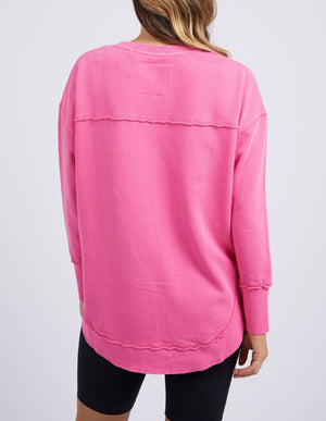 A woman with wavy hair wears a loose-fitting, long-sleeved pink Foxwood Simplified Crew made from 100% cotton with raw-edge seams and black shorts, shown from the back against a plain light background.