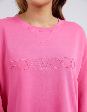 A person wears the Foxwood Simplified Crew in bright pink with "FOXWOOD" embroidered on the chest, made from 100% cotton. The image is cropped to reveal only their lower face and upper torso.