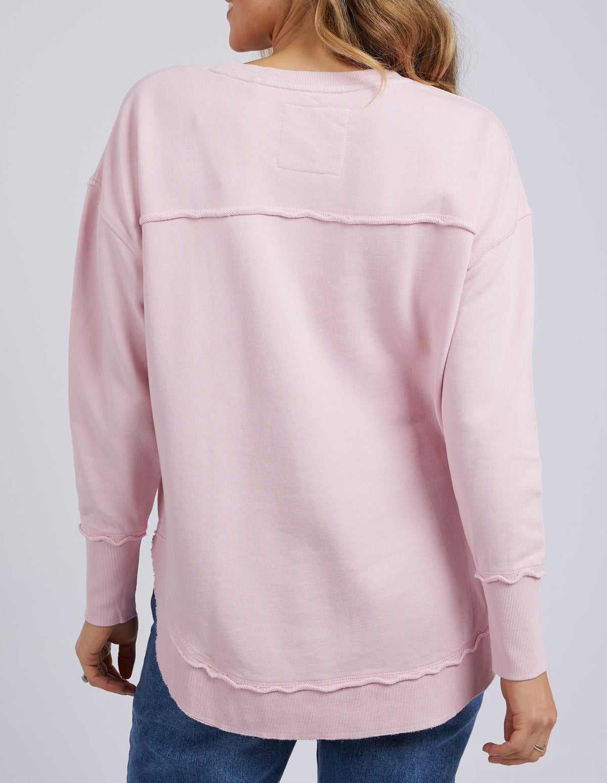 A woman with long hair, facing away from the camera, wears the Foxwood Simplified Crew—a relaxed fit light pink 100% cotton long-sleeve top with exposed seams—paired with blue jeans.